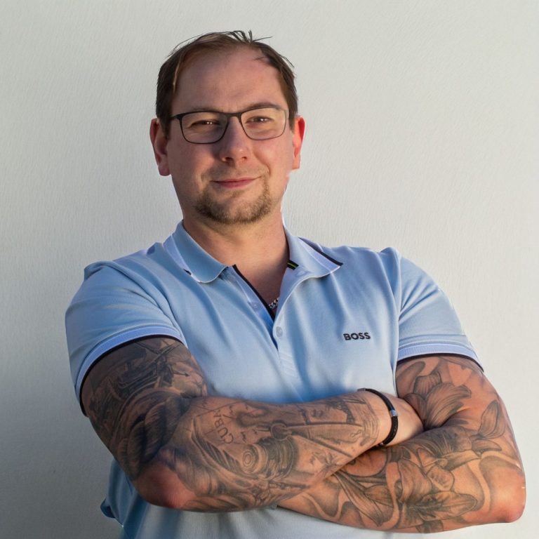 Portrait Michael Hellermann Mann mit Brille und tätowierten Armen, trägt ein hellblaues Polo-Shirt.
