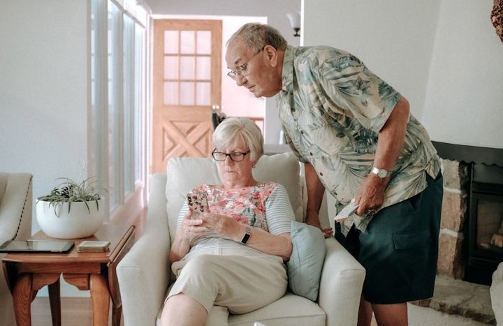 Älteres Paar sitzt zusammen, die Frau schaut auf ihr Handy, der Mann neigt sich zu ihr.