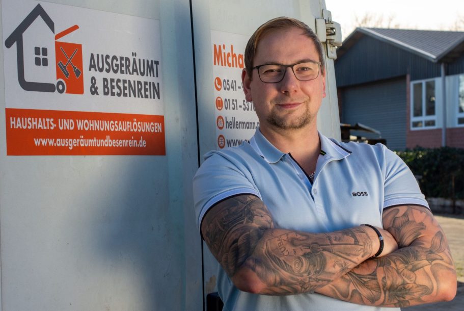 Mann mit Tattoo, lächelt und steht vor einem Fahrzeug mit einem Firmenlogo.