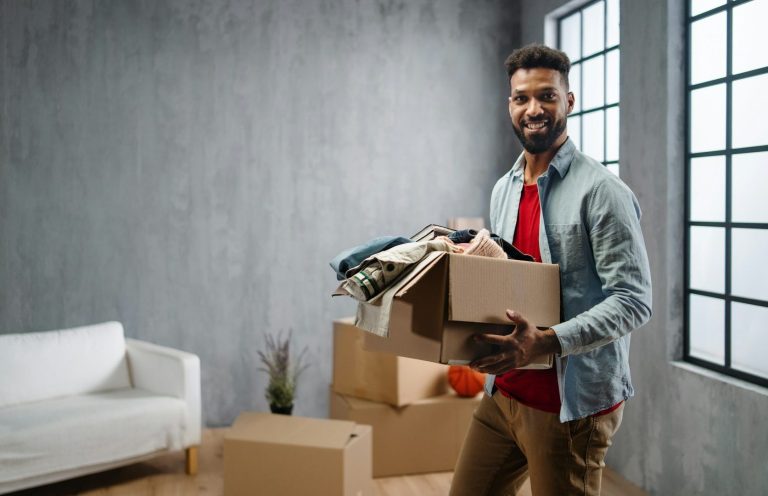 Ein Mann trägt Kisten mit Kleidung in einem Raum mit Fenstern und einer Couch.