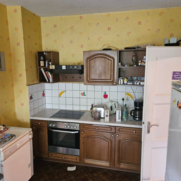 Vollgeräumte Küche mit Holzschränken, Herd, Spüle und Kühlschrank an gelber Wand.