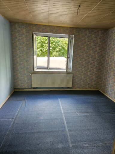 Leere Zimmeranzeige mit Fenster, hellem Teppichboden und tapetenverzierten Wänden.