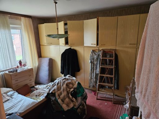 Unordentlicher Raum mit gelben Schränken, Bett und Kleidung auf dem Boden.